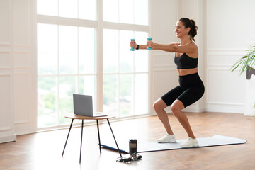 Positive fit lady exercising and squatting with two dumbbells in front of pc on coffee table at home, smiling fitness blogger recording video for her followers having personal virtual training