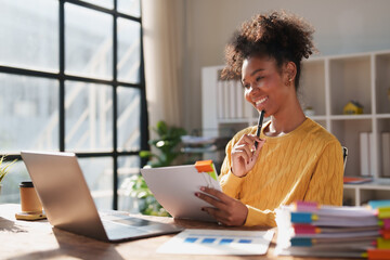 Young african american woman is smiling while holding documents and a pen, sitting at a desk with a laptop and files in a brightly lit modern office, managing documents and analyzing data