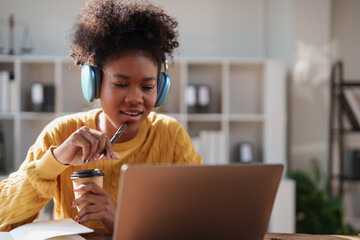 Black young woman wearing blue headphones, holding a pen and coffee cup, engaging in online learning or remote work while watching a laptop screen from a home office or study space