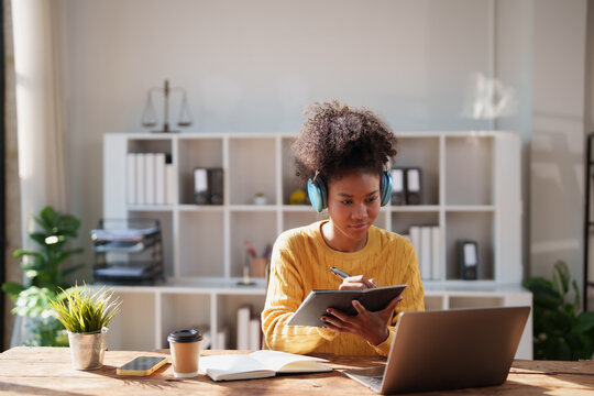 Young African American woman with afro hair wearing headphones, focused on remote learning at home, taking notes in a notebook while using a laptop for online study and productivity