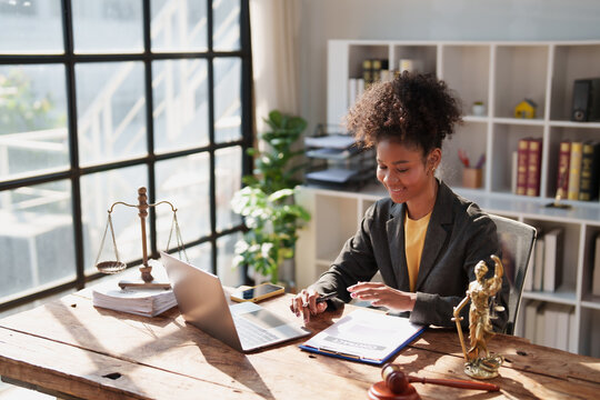 Young african american woman lawyer working on a legal contract at her desk in a contemporary law office, surrounded by legal symbols like scales of justice and a gavel