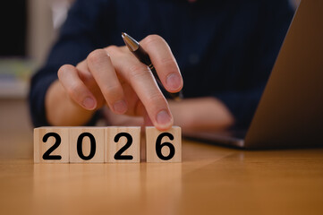 Businessman hand picks up wooden block showing the number 6 for the year 2026, Man using pen and computer laptop in modern office and showing wood block with numbers 2026