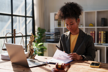 Young African American female lawyer reviewing legal paperwork, working at her desk with a laptop,...