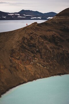 View of a solitary figure in a red jacket stands atop a craggy, brown mountain, overlooking a serene, milky blue lake under a cloudy sky, Askja, Highlands, Iceland.