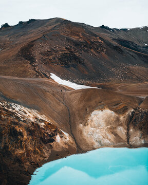 View of a turquoise lake mirroring the rugged, rust-colored terrain of a volcanic landscape under a muted sky, Askja, Highlands, Iceland.