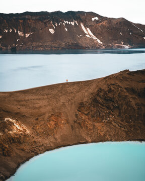 View of a solitary figure standing atop a rugged, rust-colored ridge overlooking the turquoise waters of two crater lakes in Askja, Highlands, Iceland.