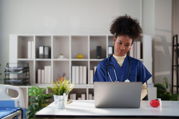 Young african american medical professional wearing scrubs and stethoscope using a laptop, focusing on patient records and healthcare data in a modern clinic setting