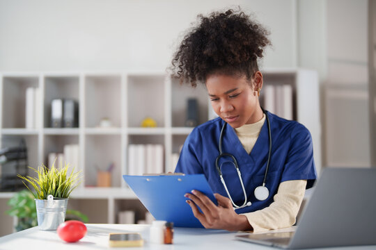 African American female nurse wearing blue scrubs and a stethoscope, intently reviewing and writing notes on a clipboard while working at a modern clinic desk with a laptop and various medical items
