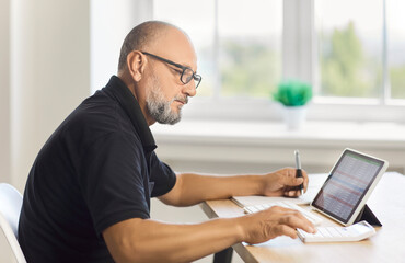 Mature man working with spreadsheet on tablet screen and calculator in office. Senior accountant sitting at desk with keyboard to review finance data and check accounting report at home workspace