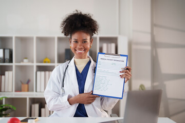 Smiling African American doctor in scrubs with stethoscope holds patient information form on a clipboard while seated at a clinic desk, offering professional care and consultation