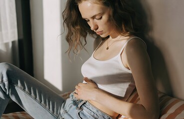 Contemplative Indoor Moment &ndash; Person in White Tank Top and Jeans Sitting in Sunlight with Hand on Stomach, contemplation, indoor, emotion, sunlight, jeans, tanktop, solitude, 