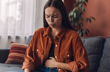Abdominal Pain Concept &ndash; Person in Rust Shirt Clutching Stomach with Pained Expression on Couch in Home Setting, stomachache, abdominal pain, digestive issue, discomfort, health, medical, couch, shirt