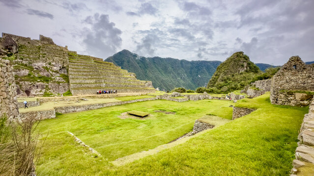 View of ancient stone structures rise amidst verdant terraces, a testament to history under a cloudy sky, framed by distant mountains, Machu Picchu, Peru.