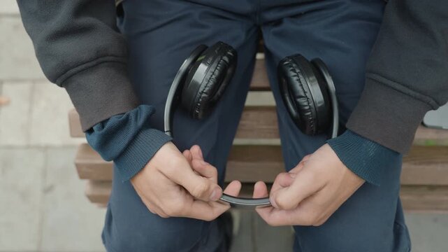 man on bench holding glasses and headphones on lap, wearing layered knit sweater and blue trousers, fingers adjusting frames in a quiet urban pause, candid closeup of hands and accessory, muted