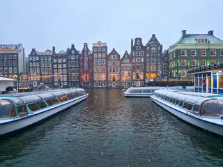 A snowy winter scene in the heart of Amsterdam's historic Damrak canal area, featuring typical narrow, gabled Dutch houses and several glass-covered canal tour boats docked in the water