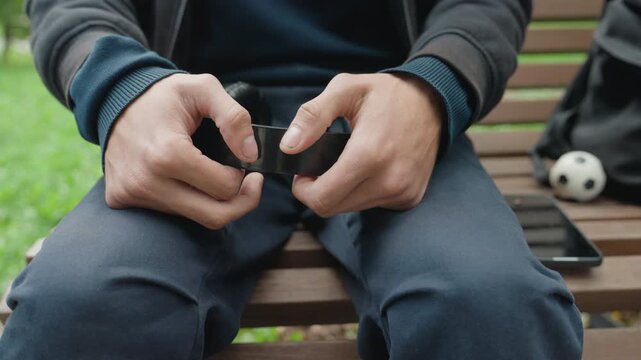 man sitting on bench holding cuff, closeup of hands unfastening black restraint, backpack and soccer ball beside seat, tablet on bench, overcast outdoor mood with tense hopeful tone