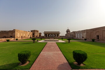 View of a symmetrical garden with manicured lawns, geometric pathways, and historic buildings under a hazy sky, Lahore, Punjab, Pakistan.