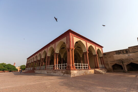 View of a building with red arches and columns stands prominently under a clear sky, birds flying above, a blend of history and open space, Lahore, Punjab, Pakistan.