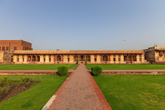 View of a brick pathway leading to an arched building with lush green lawns and manicured bushes under a clear blue sky, Lahore, Punjab, Pakistan.