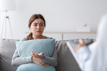 Sad unhappy upset european young female sitting on sofa hugging pillow and talking to psychologist...