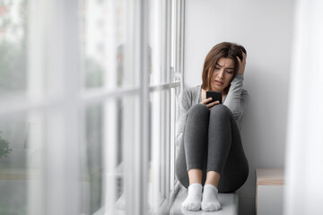 Despaired upset millennial millennial woman sitting on windowsill at home and reading message on...