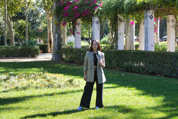 A young, attractive Spanish woman with dark hair walks around and strikes different poses in the park. She is wearing black jeans, a black T-shirt with a scarf around her neck, and a beige blazer.