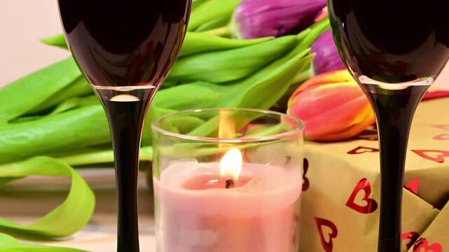 a glass of red wine to reveal a romantic tableau featuring a second glass, a lit candle, a heart-patterned gift box, and fresh tulips.