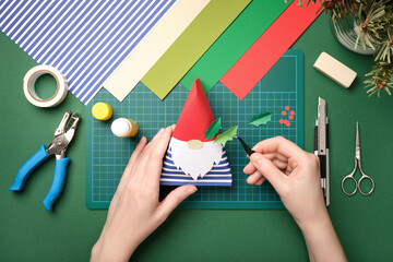 Hands assembling a DIY Christmas gnome from colored paper on a green cutting mat. Flat lay of handmade holiday decorations, creative hobby