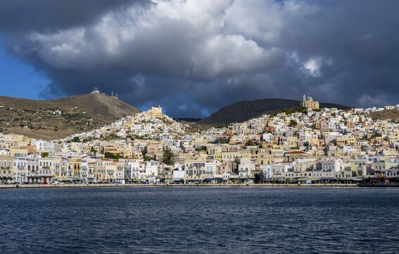 View of the town of Ermoupoli with pastel-coloured houses and sea, on the hill Anastasi Church or Church of the Resurrection and Basilica of San Giorgio in Ano Syros, dramatic cloudy sky, Ermoupoli, Syros, Cyclades, Greece