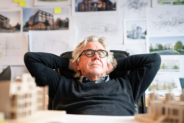 A relaxed man in a black sweater rests in an office chair, surrounded by architectural designs and a model building on his desk.