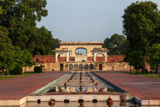 View of the Baradari in the Hazuri Bagh, with its weathered sandstone facade reflecting in the still water, framed by lush trees, Lahore, Punjab, Pakistan.
