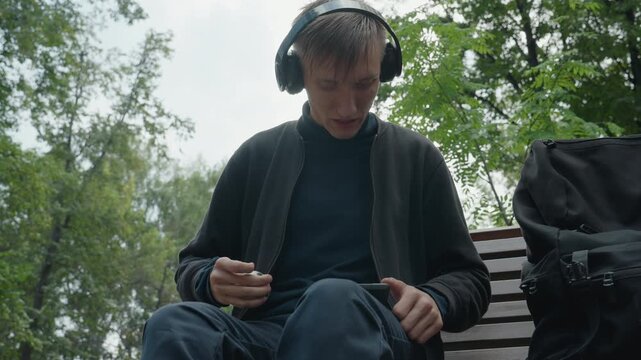 young white man on bench inspecting small spherical gadget with headphones on, closeup of hands holding toy or fidget device, backpack beside him, overcast trees, careful concentration, tactile