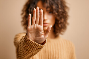 Woman with curly hair wearing a cozy brown sweater holding up her hand in a stop gesture with blurred neutral background expressing refusal or caution