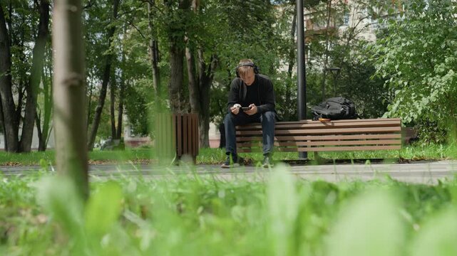 actor sitting on park bench, rehearsing lines with focused expression, earphones in, backpack at side, grass foreground, leafy trees, ambient city sounds, patient determination, streetlevel