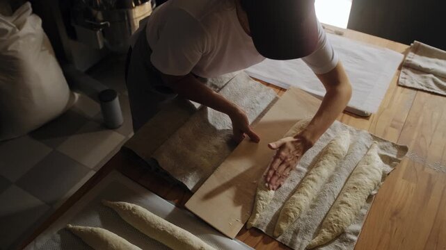 An artisan baker carefully places uncooked baguettes on a baking sheet before baking&mdash;a traditional bread-making process in an artisan bakery kitchen. Small bakery, handmade bread, morning baking. 