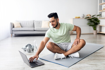Handsome Arab man sitting in lotus pose in front of laptop, working out to web sports tutorial at...