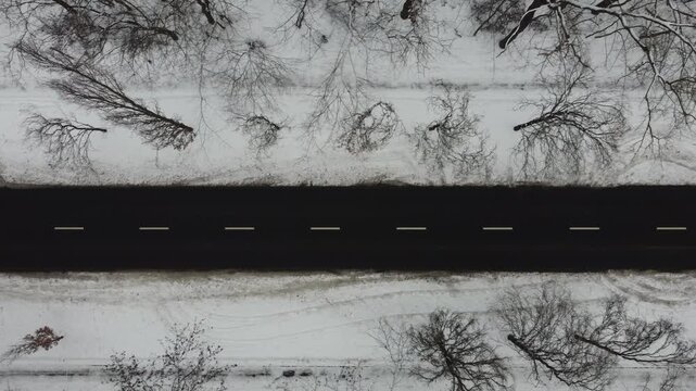 Cinematic drone footage of a snowy winter road seen from a top down aerial view. Empty asphalt highway running through frozen forest landscape with bare trees and pine woodland. Cold weather, frost.