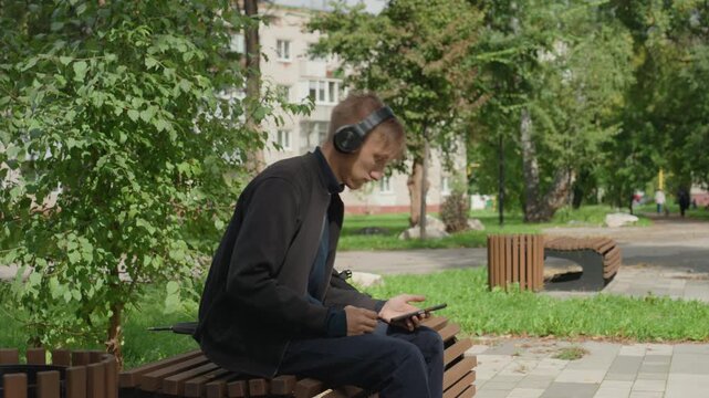 white man slouched on bench headphones, checking smartphone with downward gaze, city buildings in background, shaded tree canopy, subdued color palette, solitary commuter waiting, tired posture,