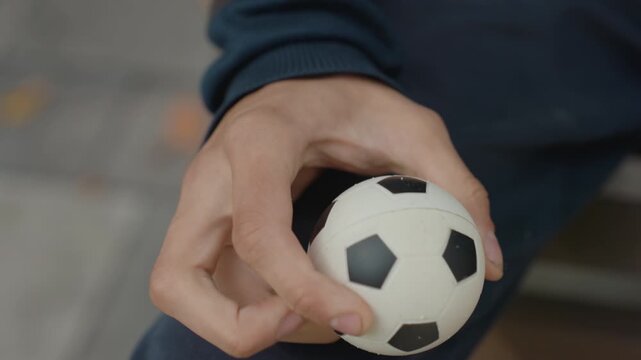closeup man hand squeezing foam soccer ball, seated on bench wearing navy sleeve, focused slow grip repetitions for hand strength and rehabilitation, textured black pentagon pattern visible, calm