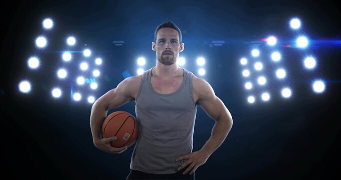 Holding orange basketball, male athlete posing at gym with gray tank, LED panels, lens flare