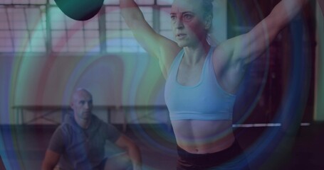Lifting woman in sports bra and dark leggings pressing kettlebell overhead in gym, coach observing