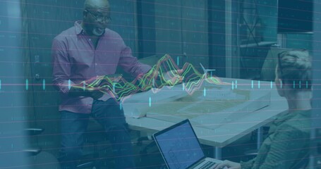 Talking pink-shirt man and blazer-clad woman leaning over office table with plans laptop and graphs