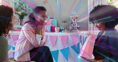 Opening pink gift bag, right woman smiling while two friends reacting at home table, with cupcakes