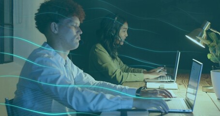 Typing coworkers wearing button-up and olive blouse at shared desk, laptops, headsets, teal wave
