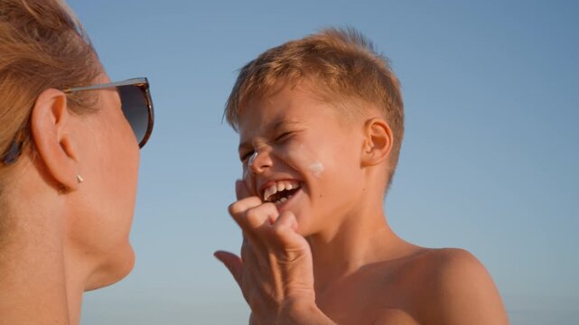 Close up of a mother applying sunblock cream on her smiling son's face during a summer holiday on a sunny beach. Health and skin protection concept.