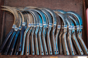 High angle view of a pile of metal sickles or reaping hooks stacked on a wooden surface. The agricultural tools feature curved blades, blue painted handles, and white stickers indicating the price.