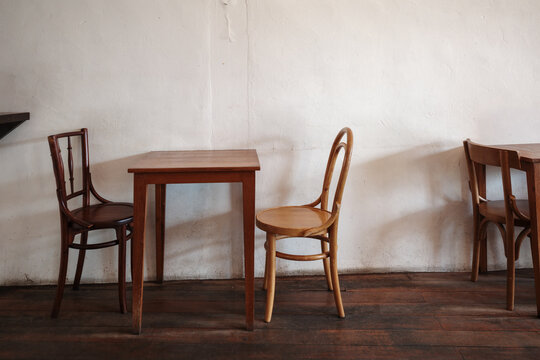 Interior view of a rustic room featuring vintage wooden bentwood chairs and square tables arranged against a textured white wall on a dark wooden floor. 