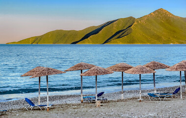 straw umbrella and sun loungers on a rocky beach in front of blue sea  and green mountain  background in Peloponnese Greece