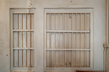 Front view of two vintage window frames embedded in a textured white wall. The windows are secured with painted vertical bars and latches, resembling a traditional or prison-style enclosure.