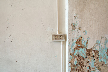 A close-up view of a weathered interior wall featuring a surface-mounted electrical outlet and conduit pipe. The adjacent corner displays severe decay with peeling beige paint.